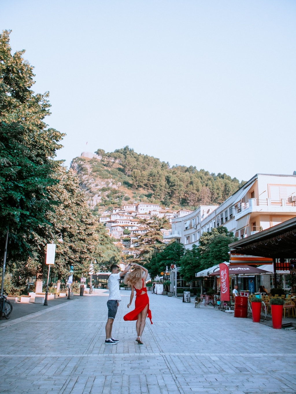 A photo of a couple twirling in the middle of the street in Berat. SHe is wearing a long, bright red dress, while he is wearing blue shorts and a white shirt. There are shops on the right with the left side lined by trees. There's a big pointed hill in the background, with the white and orange Ottoman buildings leading up the hill.