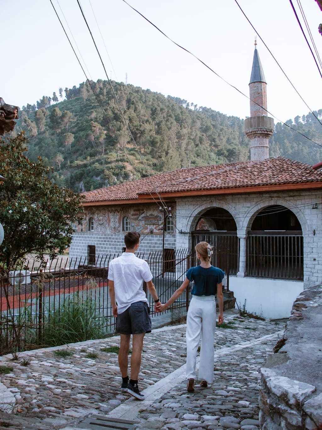 A photo of the King Mosque in Berat, Albania, with a couple walking towards it, hand in hand. The Mosque has an orange roof with a pointy minaret made of grey stone. The walls have two big archways in.