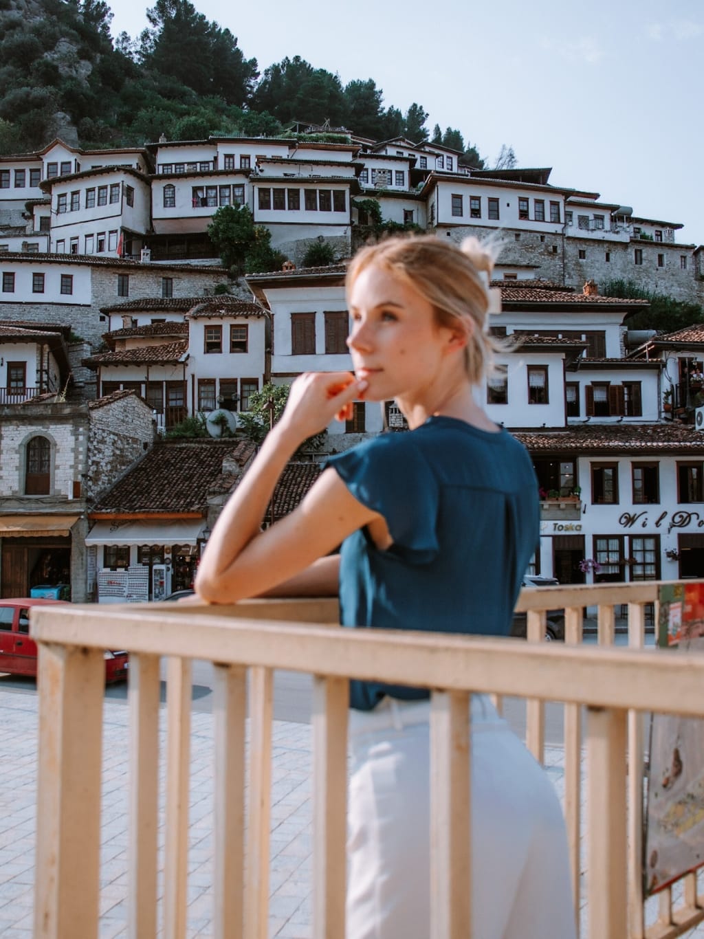 A photo of a woman wearing a blue blouse and white trousers, overlooking the beautiful Mangalem district, with it's white stone walls and orange roofs climbing up the hill behind