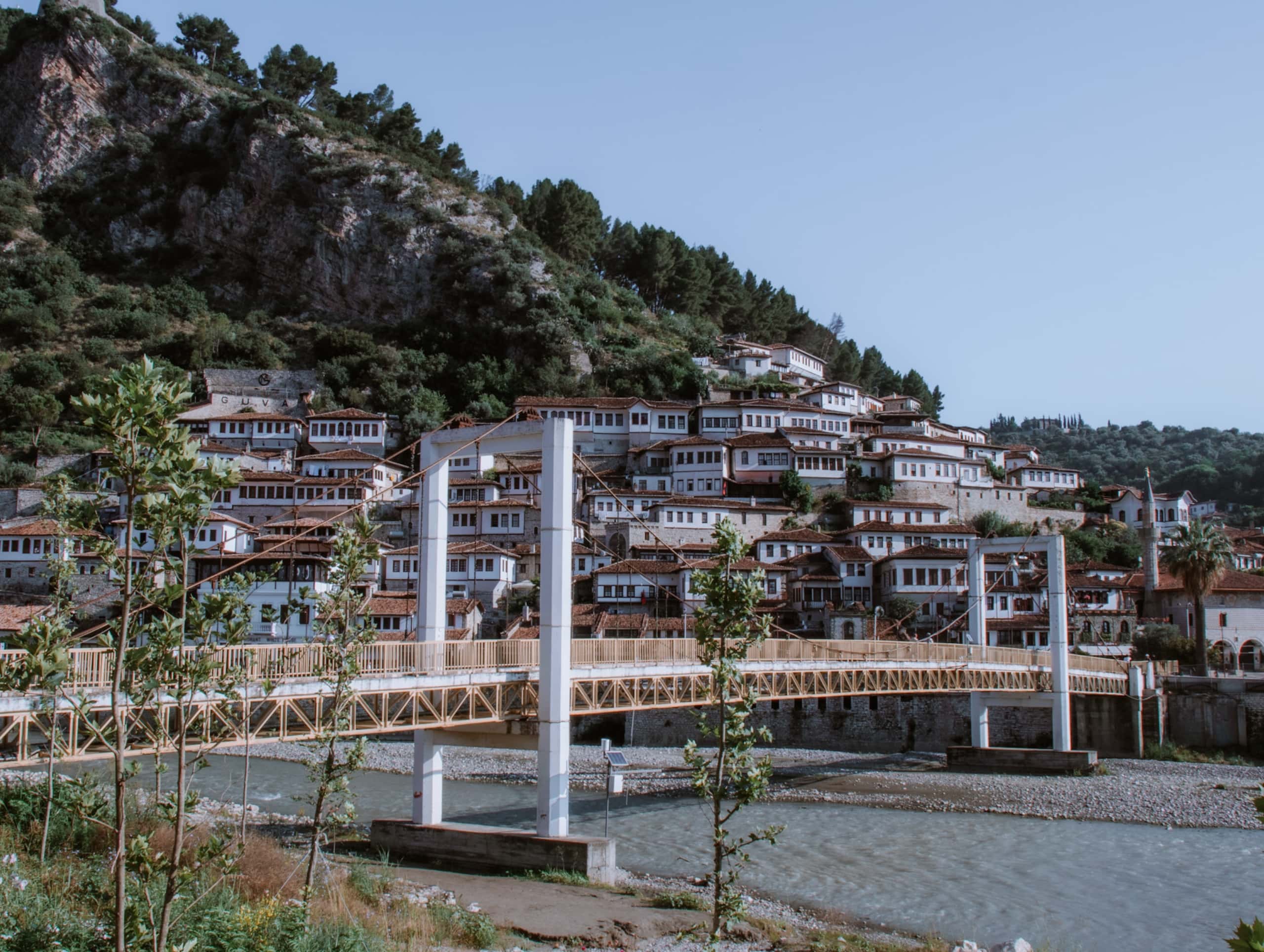 A photo showing a suspension bridge over a blue river in Berat with the Mangalem quarter seen behind. The buildings in the mangalem quarter all have orange roofs, white walls, and hundreds of rectangular windows can be seen. Behind the buildings is a hill leading up to the left, covered in green trees