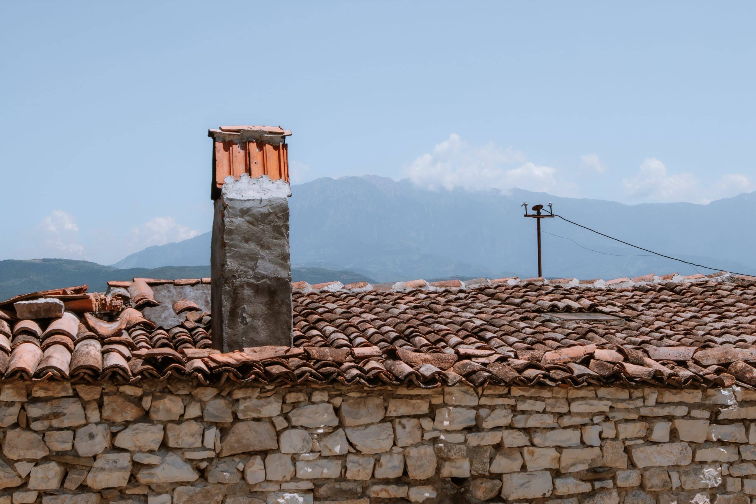 A photo showing the orange tiled roof of a stone house with a stone chimney on the left and mountains in the background