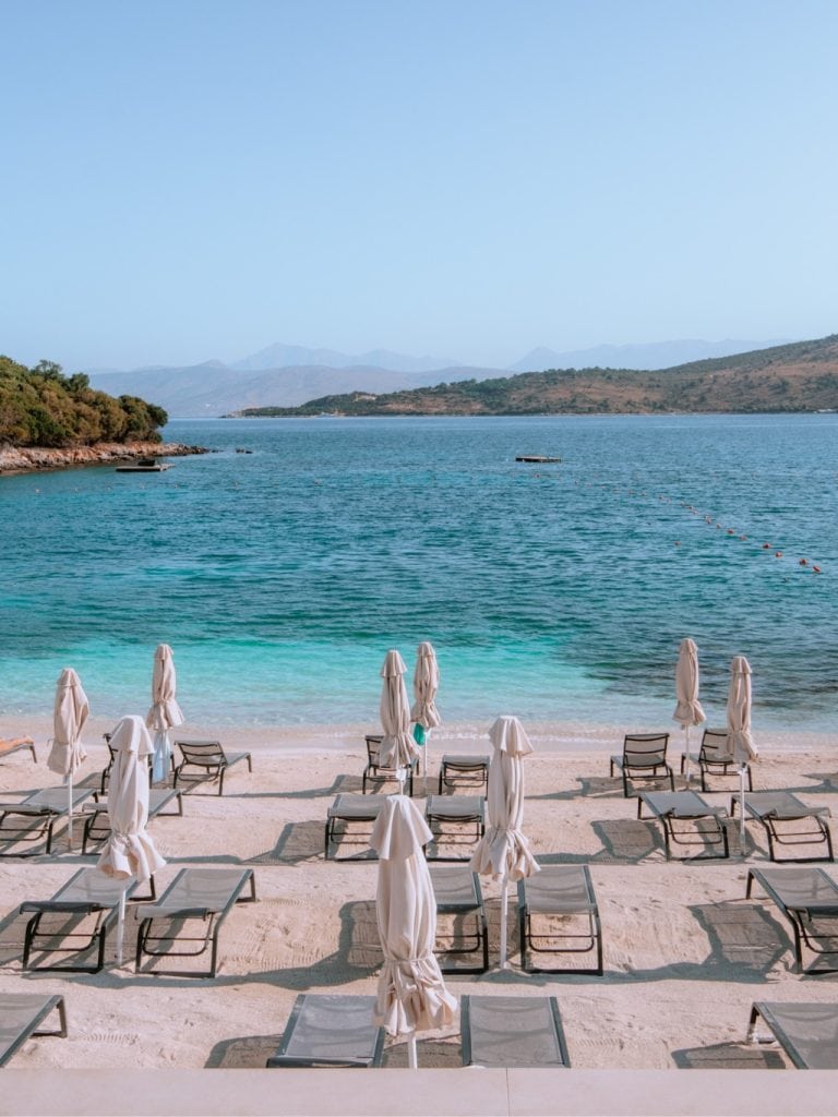 A photo of a beach in Ksamil showing white sand, and pairs of black sunloungers laid flat, with a white umbrella between, still tied down. The sea is turqoise and theres a small island visible on the left, with an outcrop of land in the distance on the right