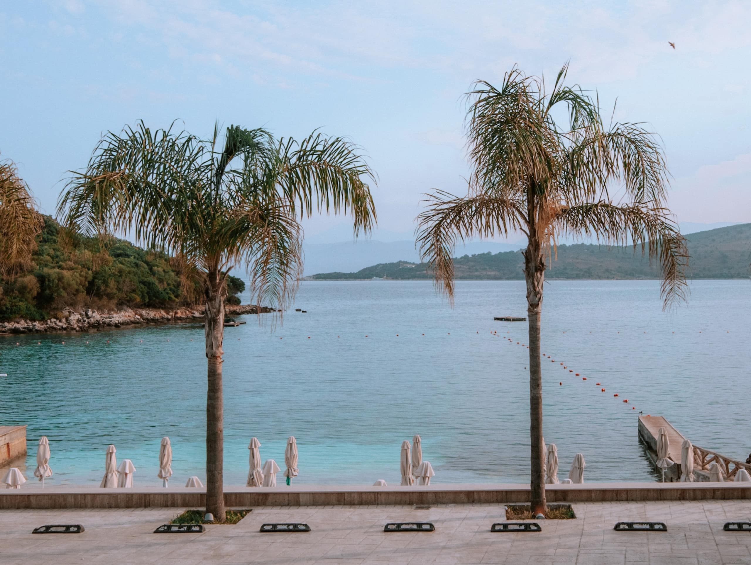 A photo of the beachfront in Ksamil. There are tow palm trees with bright blue water stretching to two islands.