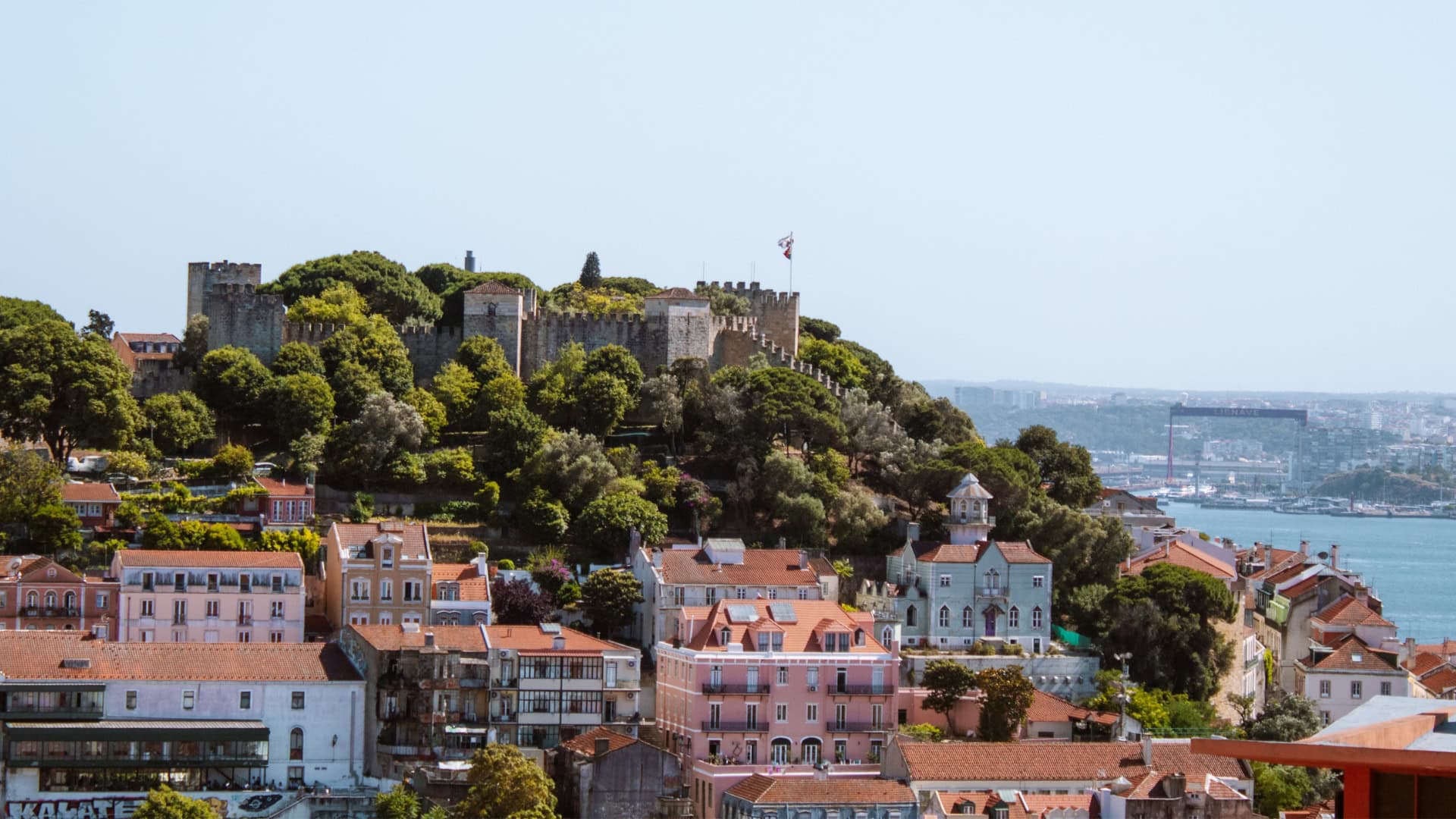 A photo of São Jorge Castle in Lisbon, perched on top of a hill, surrounded by trees, with colourful buildings below
