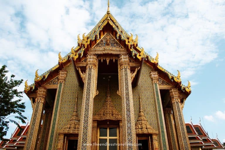 Ornate facade of a Thai temple with gold trim and intricate carvings, set against a blue sky with scattered clouds.