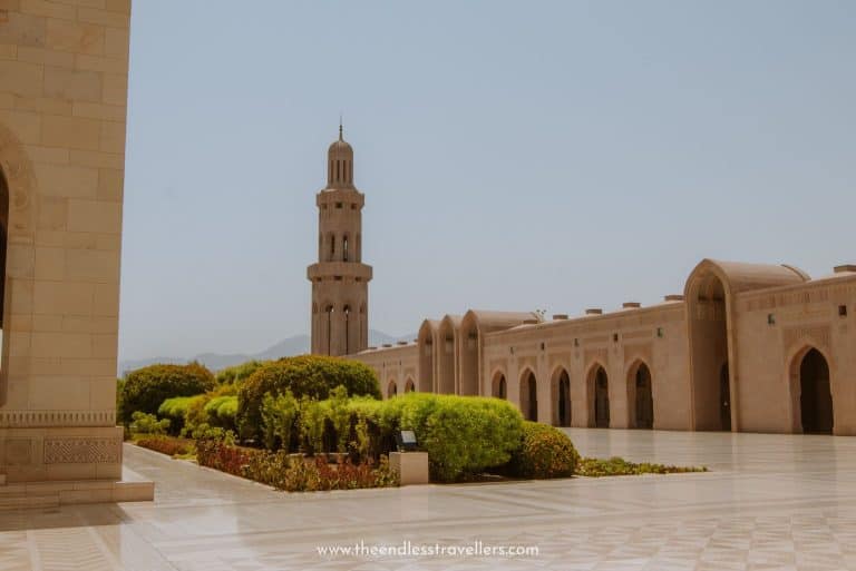 Courtyard view of Sultan Qaboos Grand Mosque in Muscat, with a tall sandstone minaret rising beside manicured gardens and arched colonnades under a clear blue sky.