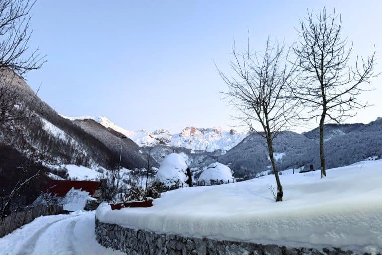A snow-covered mountain village in Albania with red-roofed houses, bare trees, and the Accursed Mountains glowing in the background at dusk. This scenic winter view helps answer the question does it snow in Albania.