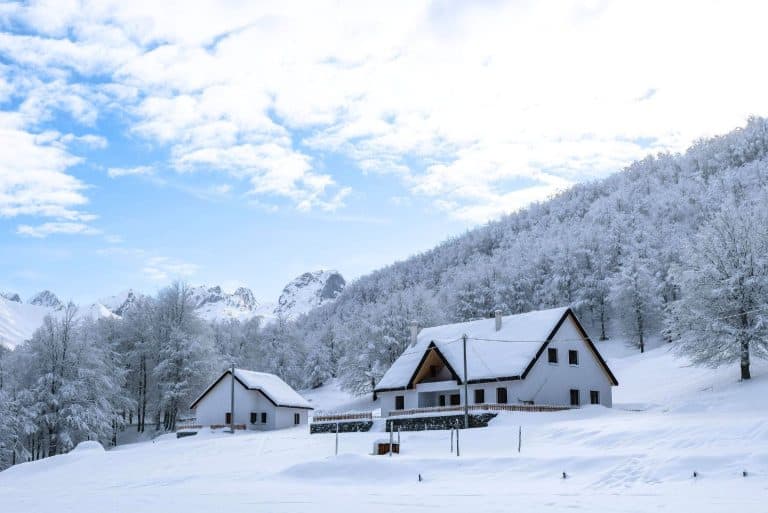 Two snow-covered alpine cottages nestled among frosty trees and mountainous terrain, under a bright blue sky. This idyllic winter scene represents remote highland living in Albania’s colder months.