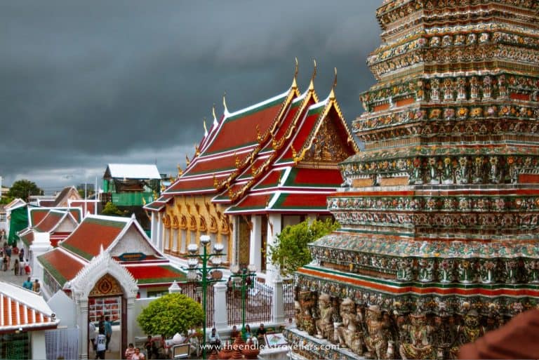 Ornate Thai temple roofs and a richly decorated stupa at Wat Arun under a dark, rainy-season sky in Bangkok, with tourists walking below.