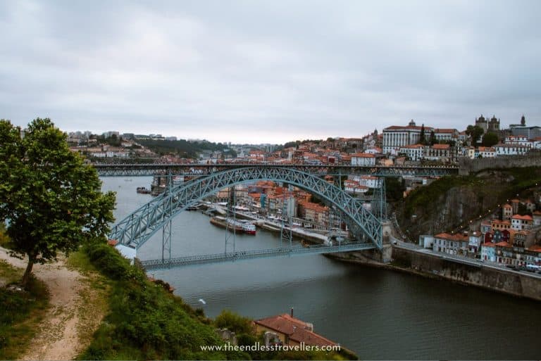 View of the Dom Luís I Bridge spanning the Douro River in Porto under a moody winter sky, with colorful riverside buildings and boats lining the waterfront.