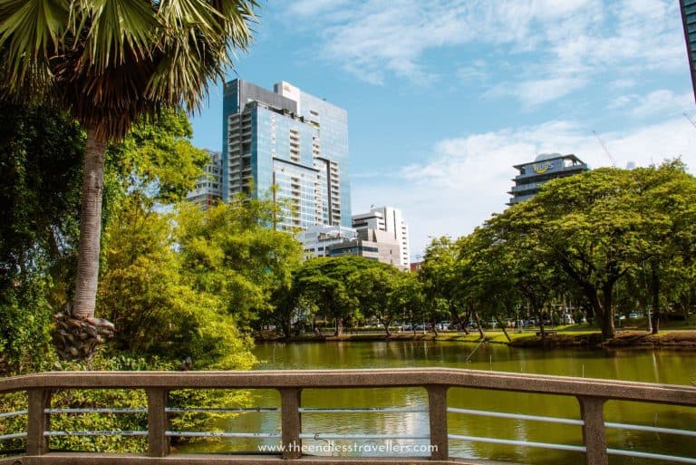 View from a footbridge in Lumphini Park showing a tranquil lake, lush greenery, and high-rise buildings in the background representing modern Bangkok.