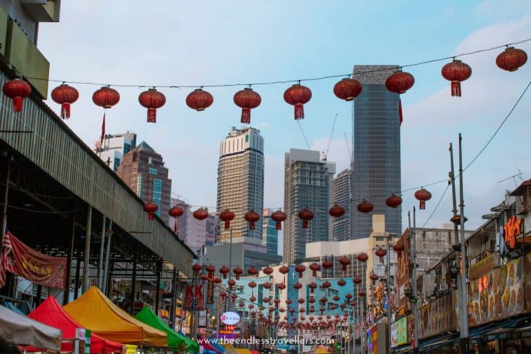 Lanterns strung above the bustling streets of Kuala Lumpur’s Chinatown with colorful market stalls below and skyscrapers rising in the distance, making this cultural hub one of the best things to do in Kuala Lumpur.