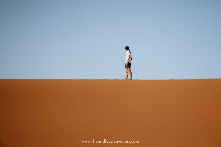 A man in a white shirt and headscarf stands alone atop a smooth sand dune under a clear blue sky, capturing the vastness and quiet beauty of Morocco in spring.