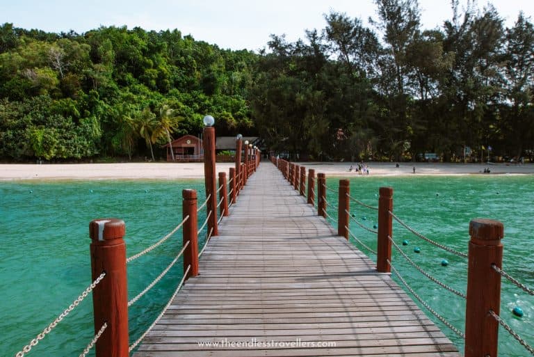 Wooden pier with red posts and rope railings stretches across bright turquoise water toward a tropical beach backed by forest. Small buildings and palm trees sit along the shoreline.